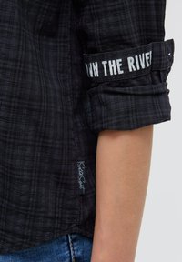 Schwarzes kariertes Hemd mit hochgekrempelten Ärmeln. Mit weißem Schriftzug "RUN THE RIVER" am Bund. Etikett am Saum mit Markennamen sichtbar.