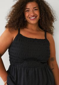 Zwarte mouwloze top met smockdetails over de bodice, met dunne bandjes en een vloeiende onderkant. Huidkleur en tatoeage zichtbaar.