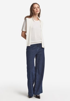 Haut à manches courtes blanc superposé avec un gilet sans manches crème, associé à un jean large bleu foncé et des escarpins noirs à bout pointu.