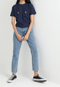 T-shirt in cotone navy con due design di ananas ricamati, abbinato a jeans denim azzurri e sneakers nere, in piedi su uno sfondo bianco.