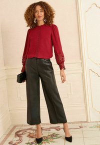Frau mit lockigem Haar, die eine rote Spitzenbluse, schwarze weite Lederhosen und schwarze High Heels trägt, während sie in einem prunkvollen Raum eine schwarze Clutch hält.