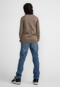 Brauner gestrickter Pullover, blaue Jeans, schwarze Sneakers mit weißen Akzenten, von hinten vor einem schlichten Hintergrund betrachtet.