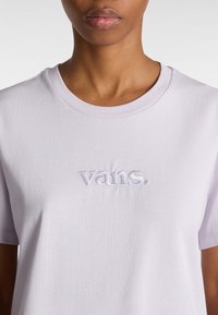 Gros plan sur une personne portant un t-shirt Vans de couleur mauve clair avec un logo brodé centré sur la poitrine, sur un fond uni.