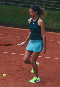 Weibliche Tennisspielerin in blauer Ausrüstung und neonfarbenen Schuhen, die einen Ball mit einem Wilson-Schläger auf einem Sandplatz aufspringt.