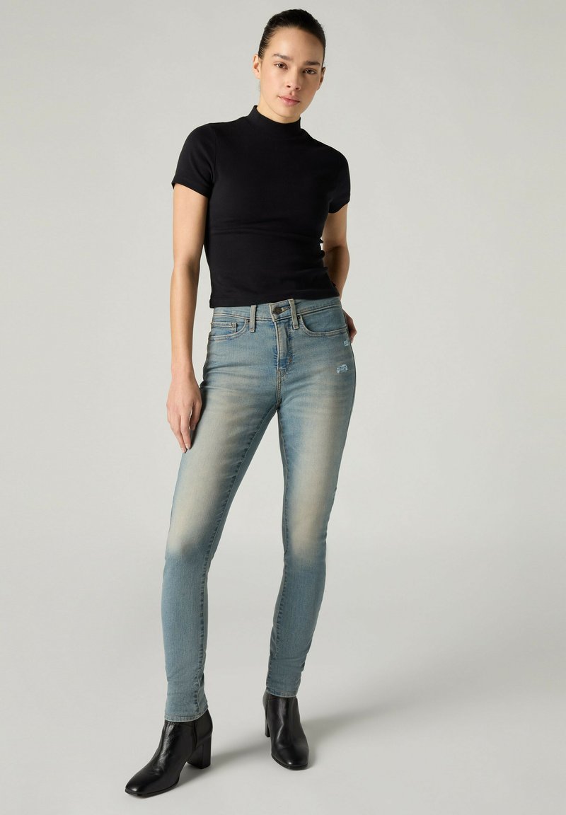 Kvinde står iført en sort top med korte ærmer, falmede blå skinny jeans og sorte ankelstøvler mod en ensfarvet lys baggrund.