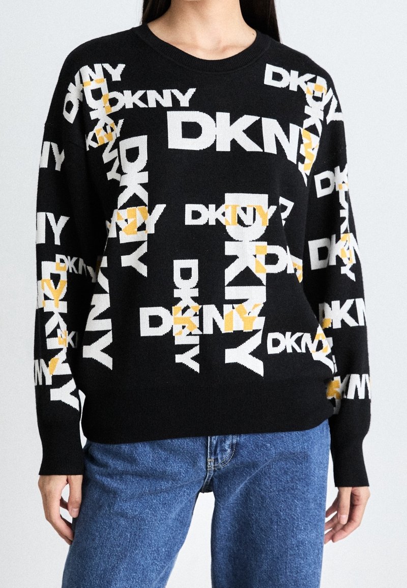 Svart tröja med rund hals och långa ärmar, med ett upprepat DKNY-logotyp i vitt och gult. Kombinerad med blått denimjeans.