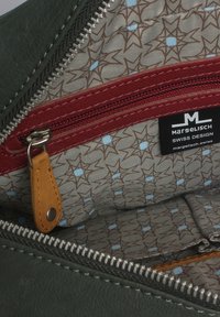 Grünes Lederhandtascheninnere, das eine rote Reißverschlusstasche, ein beiges Innenfutter mit einem Sternmuster in Braun und Hellblau sowie einem Markenetikett zeigt.