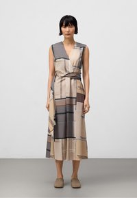 Robe portefeuille beige avec motif à carreaux géométriques ; sans manches ; tissu texturé ; lien à la taille ; longueur au genou. Chaussures marron à enfiler.