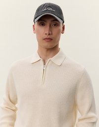 Maglione a coste crema con colletto a metà zip. Berretto grigio con testo ricamato bianco. Texture liscia e design aderente. Sfondo neutro.