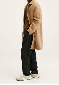 Lichtbruine wollen jas over een bruine coltrui, gecombineerd met zwarte broek en beige sneakers met witte accenten. Effen achtergrond.