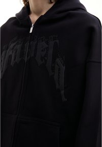 Personne portant un sweatshirt à zip noir avec un texte en relief de style gothique et de petites clous décoratifs sur la poitrine.