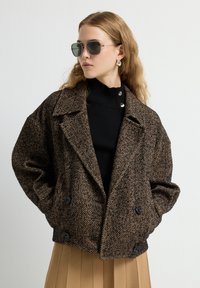 Bruine herringbone gemusterde dubbelzijdige jas met grote revers, zwarte knopen en een oversized silhouet, gedragen over een zwarte coltrui.