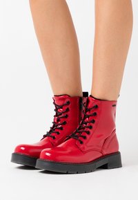 Botas de tobillo de cuero rojo brillante con cordones y suelas negras, usadas sobre piernas desnudas contra un fondo blanco.