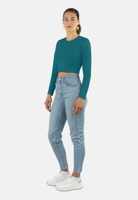 Türkisfarbener Langarm-Crop-Top, kombiniert mit hellblauen, hochgeschnittenen Jeans und weißen Sneakern. Einfaches Design, glatte Textur und taillierte Form.