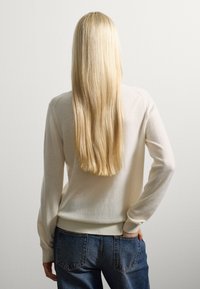 Crème-kleurige, zachte trui met lange mouwen en geribbelde zoom, gecombineerd met lichtblauwe denim jeans. Haar valt voorbij de schouders.