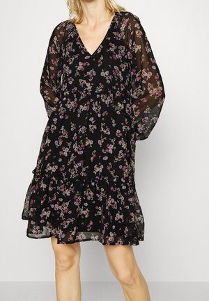 Kvinde iført en sort blomsterkjole med transparente lange ærmer og V-hals, stående mod en ensfarvet baggrund.