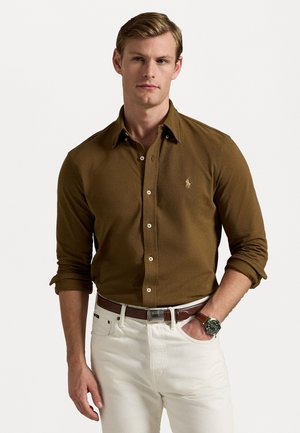 Chemise marron à manches longues avec un col classique, comportant un petit logo sur la poitrine ; associée à un pantalon blanc et une ceinture marron.