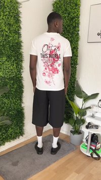Homem a usar uma camisa branca com um padrão floral rosa e o texto "Fora deste Mundo", calções pretos, meias brancas e sapatos pretos, de pé no interior.