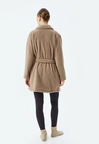 Cappotto in pelliccia sintetica marrone con colletto, vita fasciata e maniche lunghe, abbinato a leggings scuri e calzini morbidi chiari. Vista da dietro.