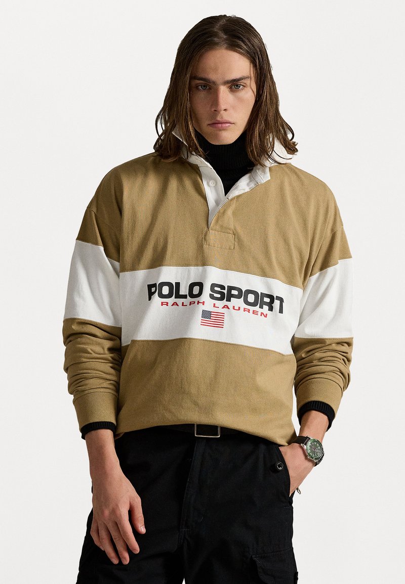 Beige en witte polo met lange mouwen en een ribbelkraag, voorzien van de tekst "POLO SPORT" en een detail van de Amerikaanse vlag. Draag het over een zwarte coltrui.