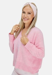 Maglione rosa chiaro e peloso con scollo a V e maniche lunghe. Il modello indossa una fascia per capelli rosa abbinata e sorride, tenendo i capelli con una mano.