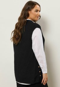 Zwarte geribbelde gebreide mouwloze vest met knoopaccenten aan de zijkant, gedragen over een witte blouse, weergegeven tegen een neutrale achtergrond.