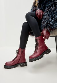 Bocanci din piele burgund cu șireturi negre, fermoar pe lateral și talpă din cauciuc texturat negru cu accente metalice pe margine.