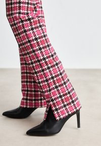 Pantalons à motif pied-de-coq rose et noir avec fentes latérales, associés à des escarpins noirs à bout pointu. Texture lisse, coupe ajustée.