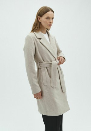 Femme portant un manteau en laine beige ceinturé avec de larges revers, debout de profil devant un fond clair uni.