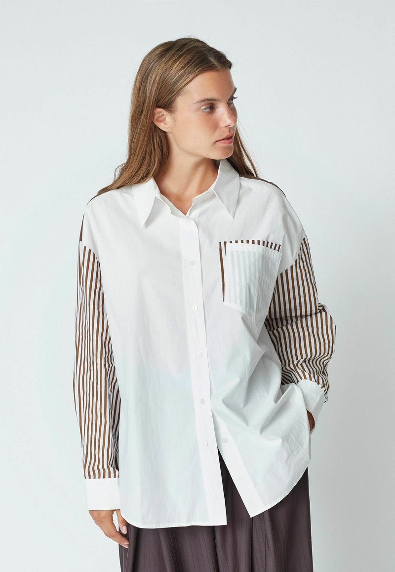 Camicia bianca con maniche lunghe a righe marroni, una tasca sul petto e chiusura con bottoni. Realizzata in un tessuto leggero con una consistenza liscia.