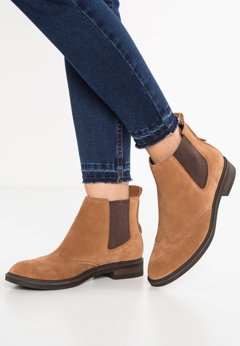 Bottes Chelsea en daim couleur noisette avec des panneaux élastiques latéraux foncés, portées avec un jean skinny bleu foncé, pieds croisés sur fond blanc.