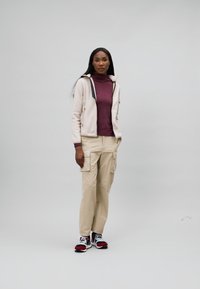 Lyserød lynlåsjakke, burgundy hultravneck, beige cargo-bukser og multicolor sneakers. Glat tekstur, moderne design med funktionelle lommer.
