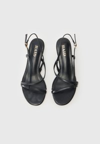 Sandal kulit hitam dengan sol datar, dilengkapi dengan tali silang tipis dan tali pergelangan kaki yang dapat disesuaikan. Nama merek "RAID" terukir di bagian dalam sandal.