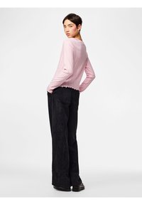 Γυναίκα με κοντά σκούρα μαλλιά, που φοράει ένα ανοιχτό ροζ μακρυμάνικο τοπ και μαύρο παντελόνι με φαρδιές φάσες, στέκεται με τα χέρια στις τσέπες, κοιτώντας πάνω από τον ώμο.