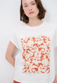 Weißes Kurzarmhemd mit einem großen orangen Blumenmuster und cremefarbenen Akzenten. Text "Zeitlose Schönheit in voller Blüte" über dem Design.