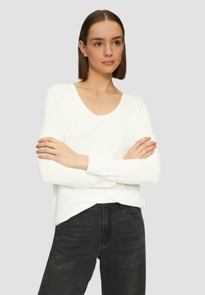 Witte lange mouw top met een V-halsontwerp, gemaakt van zachte stof. Gecombineerd met donkere jeans, met de armen gekruist voor een neutrale achtergrond.