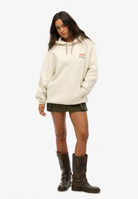 Haut à capuche couleur crème avec cordon de serrage et logo, associé à un short olive et des bottes marron jusqu'au genou. Texture plate, coupe décontractée.