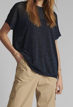 Femme portant un haut à manches courtes pailleté bleu marine avec un pantalon cargo beige, une main dans la poche, sur un fond gris uni.