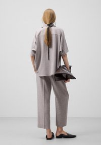 Lichtgrijze, losse shirt met een rugstrikdetail, gecombineerd met bijpassende wijde broek; houdt een bruin, glanzend clutch vast. Zwarte instappers.