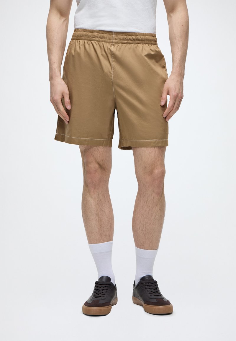 Homme portant un short marron à taille élastique, des chaussettes blanches et des baskets foncées, debout devant un fond neutre.