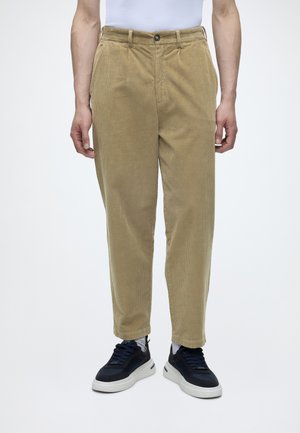 Homme portant un pantalon en velours côtelé beige, des chaussettes blanches et des baskets noires, debout devant un fond blanc uni.