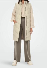 Kobieta w długim beżowym pikowanym kamizelce na jasnej koszuli, w szarych szerokich spodniach i kremowych masywnych butach, stojąca z rękami w kieszeniach.