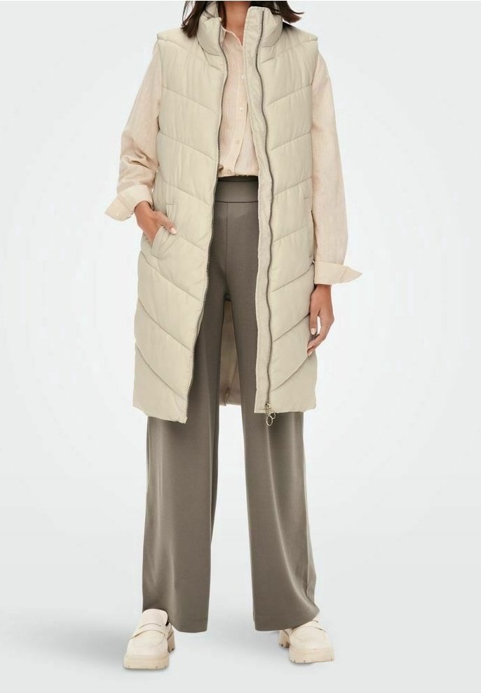 Kobieta w długim beżowym pikowanym kamizelce na jasnej koszuli, w szarych szerokich spodniach i kremowych masywnych butach, stojąca z rękami w kieszeniach.