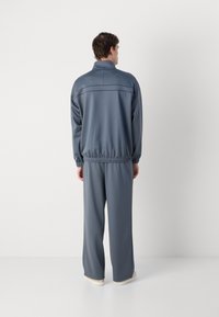 Lys grå treningsdrakt med krage, med elastiske mansjetter og midjebånd. Stoffet ser glatt ut med en avslappet passform. Hvite joggesko under.