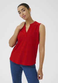 Rotes ärmelloses Oberteil mit V-Ausschnitt, versehen mit Lochmustern und einem strukturierten Stoff. Kombiniert mit blauen Jeans.