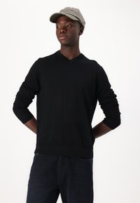 Čierny sveter s v-neck golierom a dlhými rukávmi, vyrobený z mäkkého materiálu. Dizajn obsahuje malé logo na hrudi, skombinované s tmavými džínsy a sivou šiltovkou.