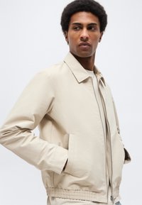 Ung mand med kort, krøllet hår, iført en beige jakke med lynlås og krave, står med hænderne i lommerne og kigger mod højre.