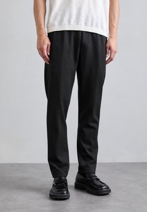 Homme portant un pantalon habillé noir, des mocassins noirs et une chemise blanche à manches courtes, debout sur un sol gris devant un fond uni clair.