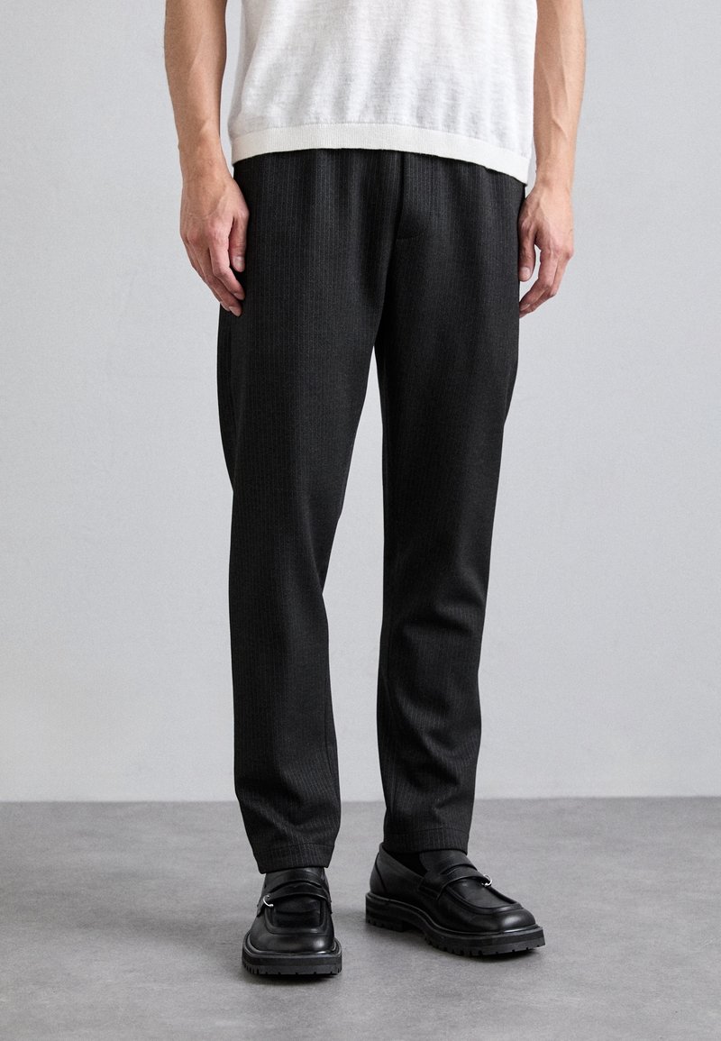 Homme portant un pantalon habillé noir, des mocassins noirs et une chemise blanche à manches courtes, debout sur un sol gris devant un fond uni clair.
