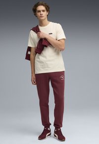 Beige T-shirt med svag logo; maroon sweatpants med logo; maroon løbesko med creme detaljer; stribet maroon sweatshirt.